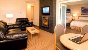 A hotel room with a fireplace, a TV mounted above it, a bed with a headboard, and a dining area with a table and chairs. - Woodlands Inn & Suites (Fort Nelson)
