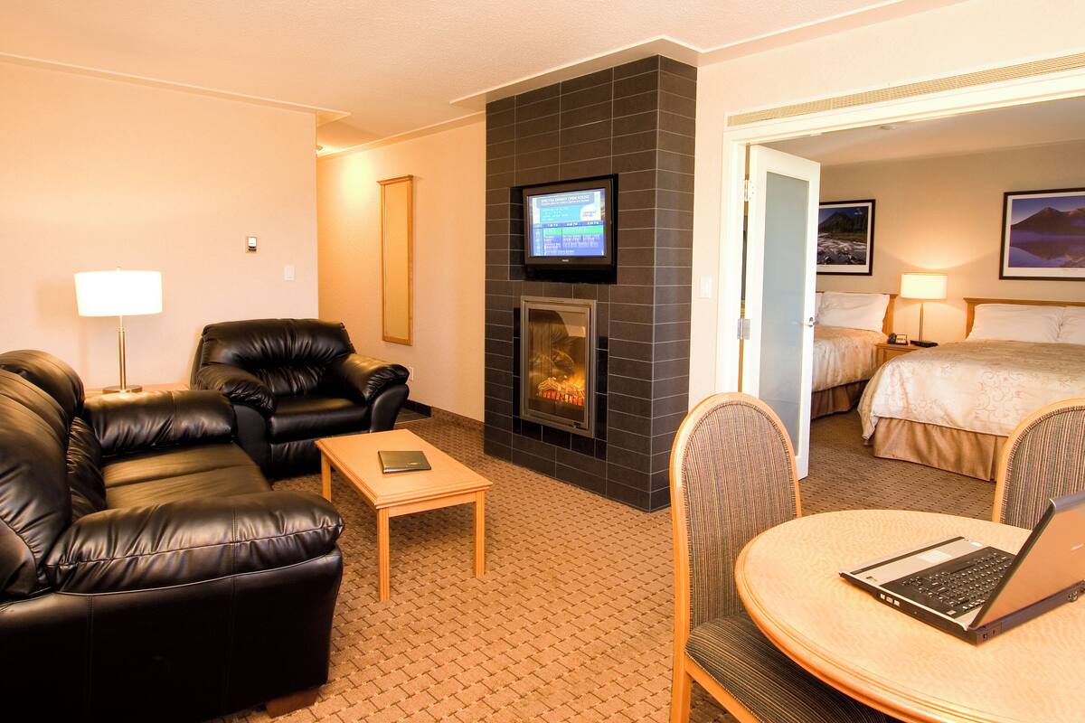 A hotel room with a fireplace, a TV mounted above it, a bed with a headboard, and a dining area with a table and chairs.