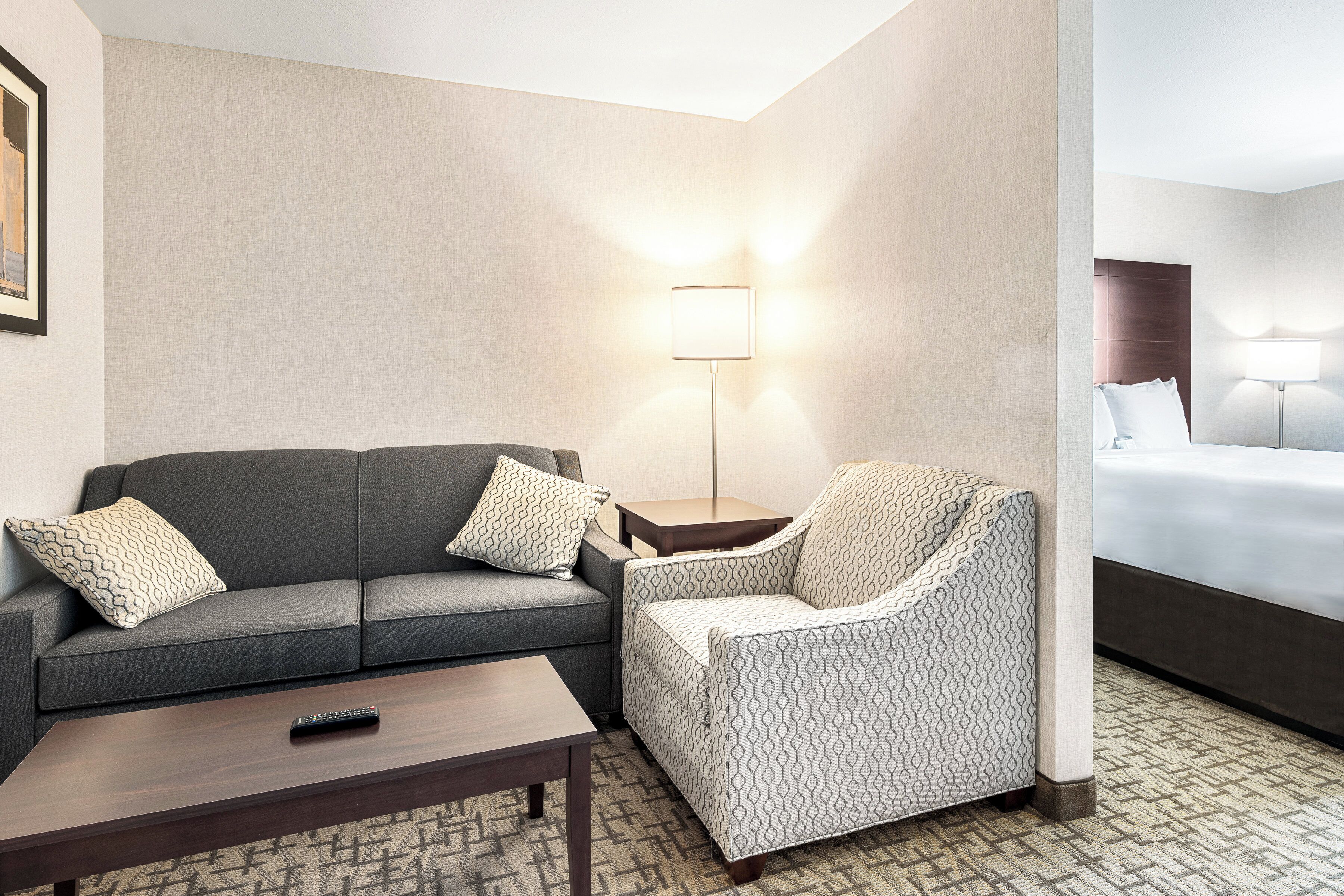 Habitación de hotel con sofá gris, sillón estampado, una mesita y una cama con cabecero.