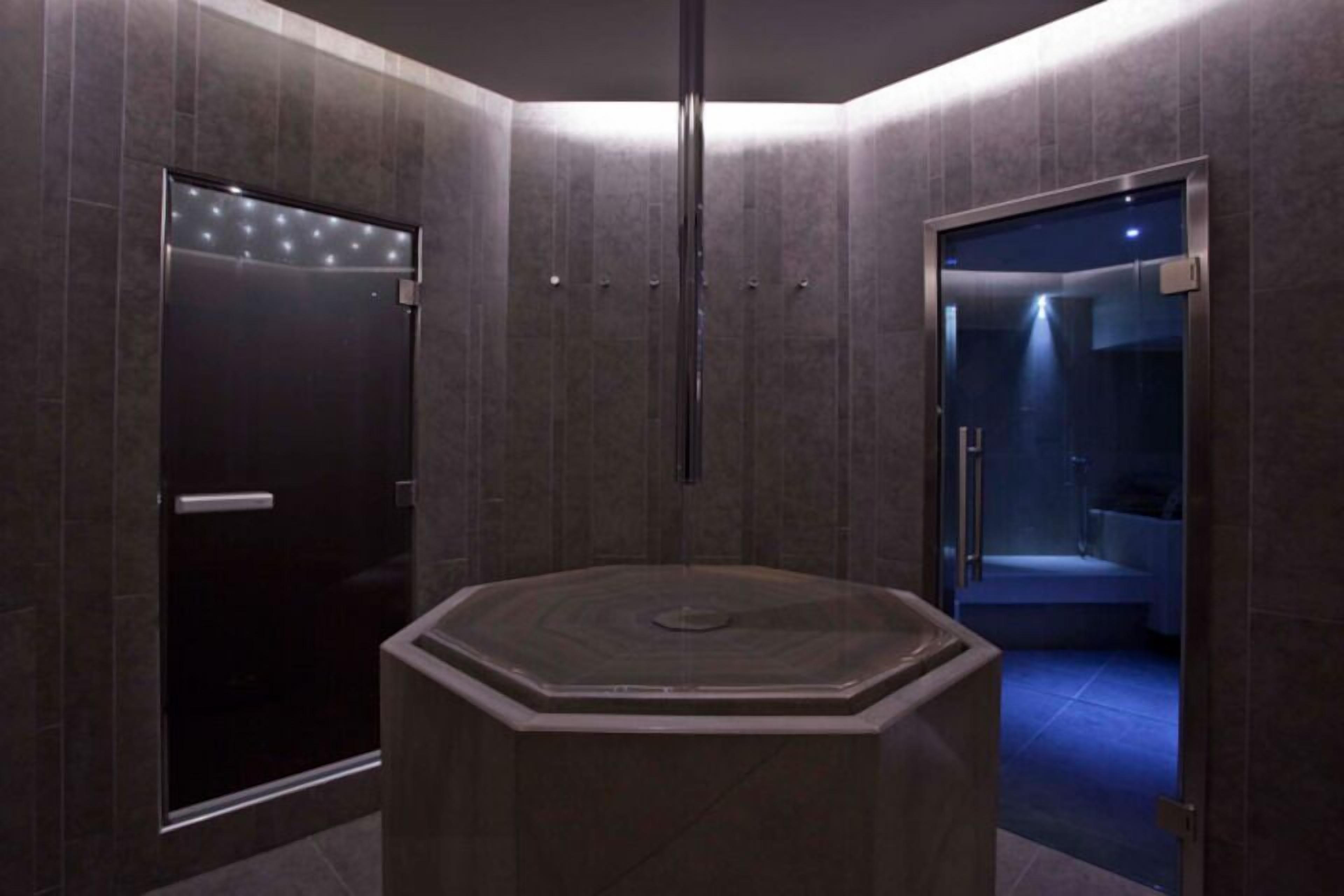A modern bathroom with a central octagonal sink, a dark door with a light strip, and a glass shower enclosure.