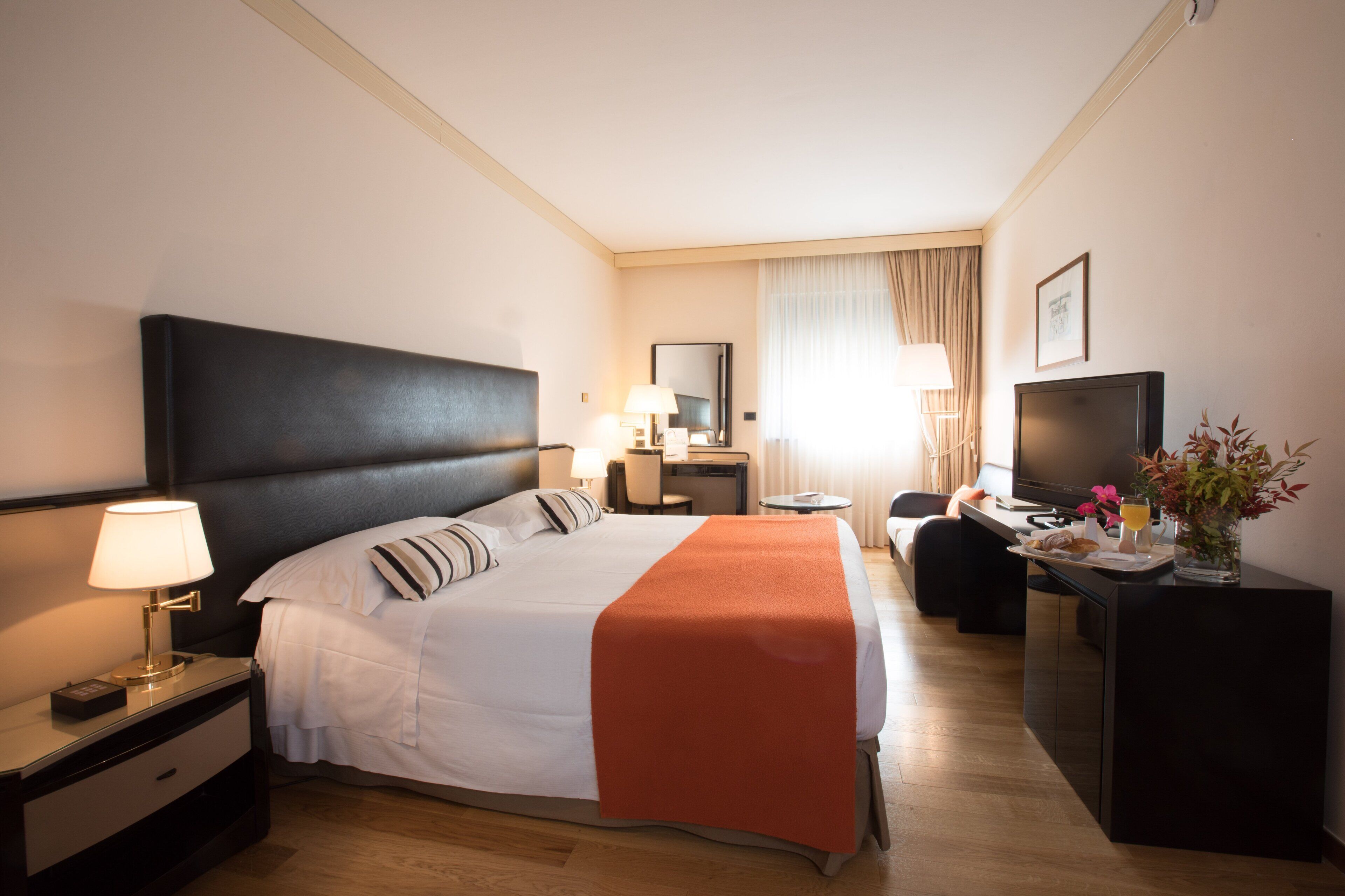 A hotel room with a large bed, a desk with a TV, a nightstand with a lamp, and a small table with a vase of flowers.