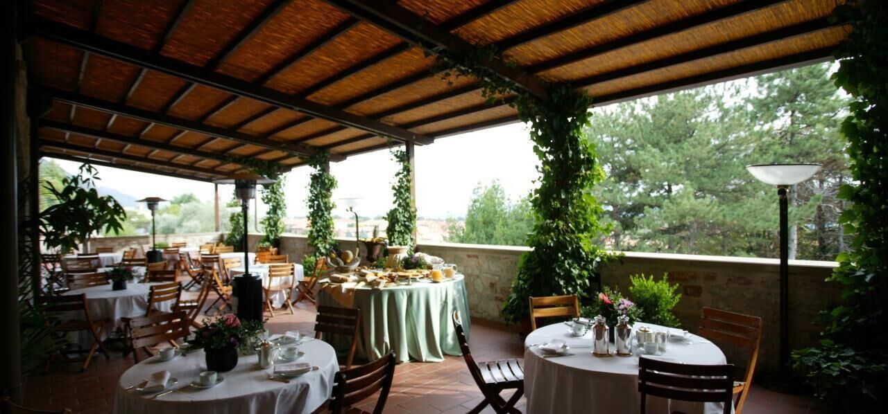 An outdoor dining area with tables and chairs, covered by a wooden roof, and surrounded by greenery.