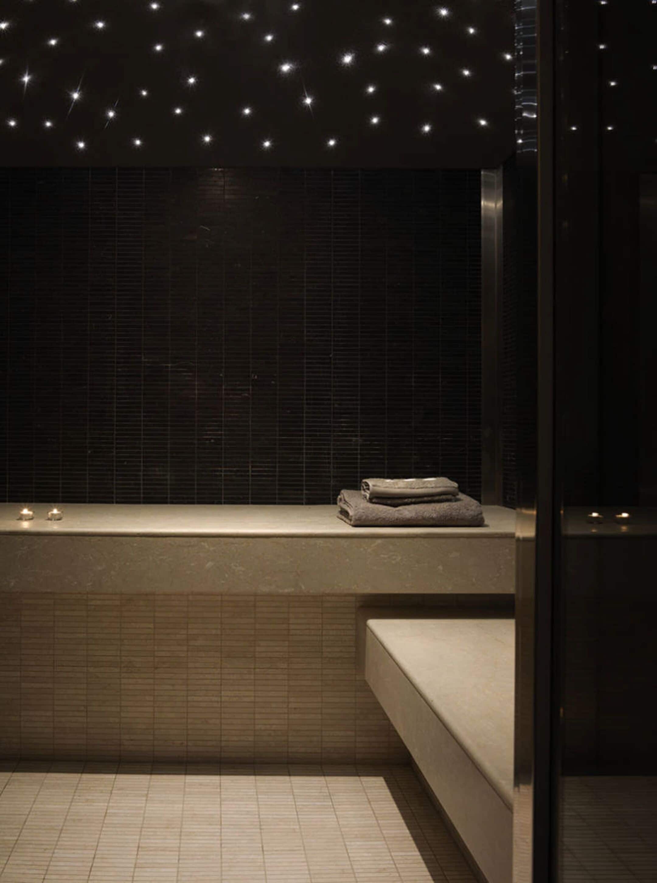 Ein modernes Badezimmer mit dunklen Fliesen, einer Sitzbank und einem kleinen Ablagebereich mit Handtüchern.