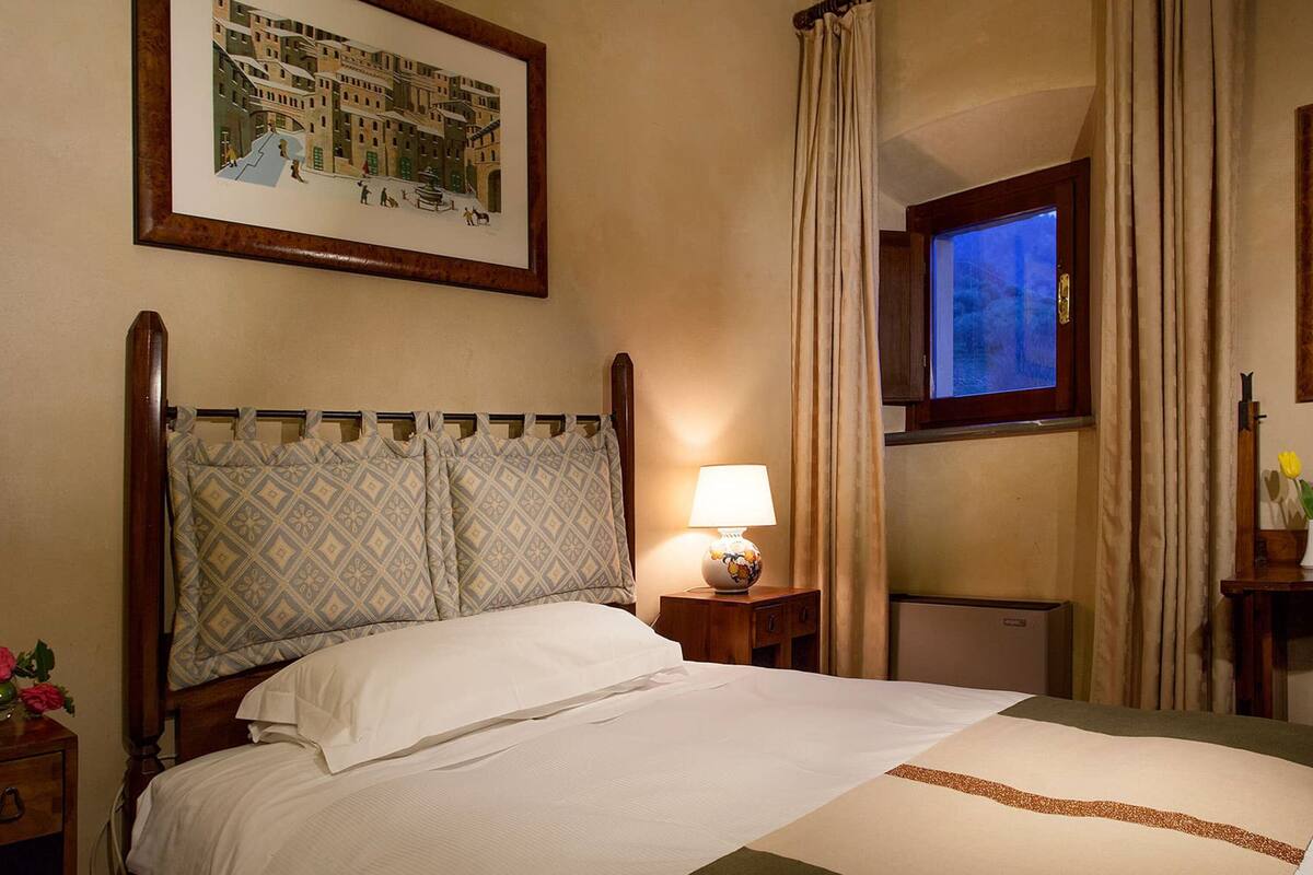 A hotel room with a bed, a nightstand with a lamp, a window with curtains, and framed pictures on the walls.