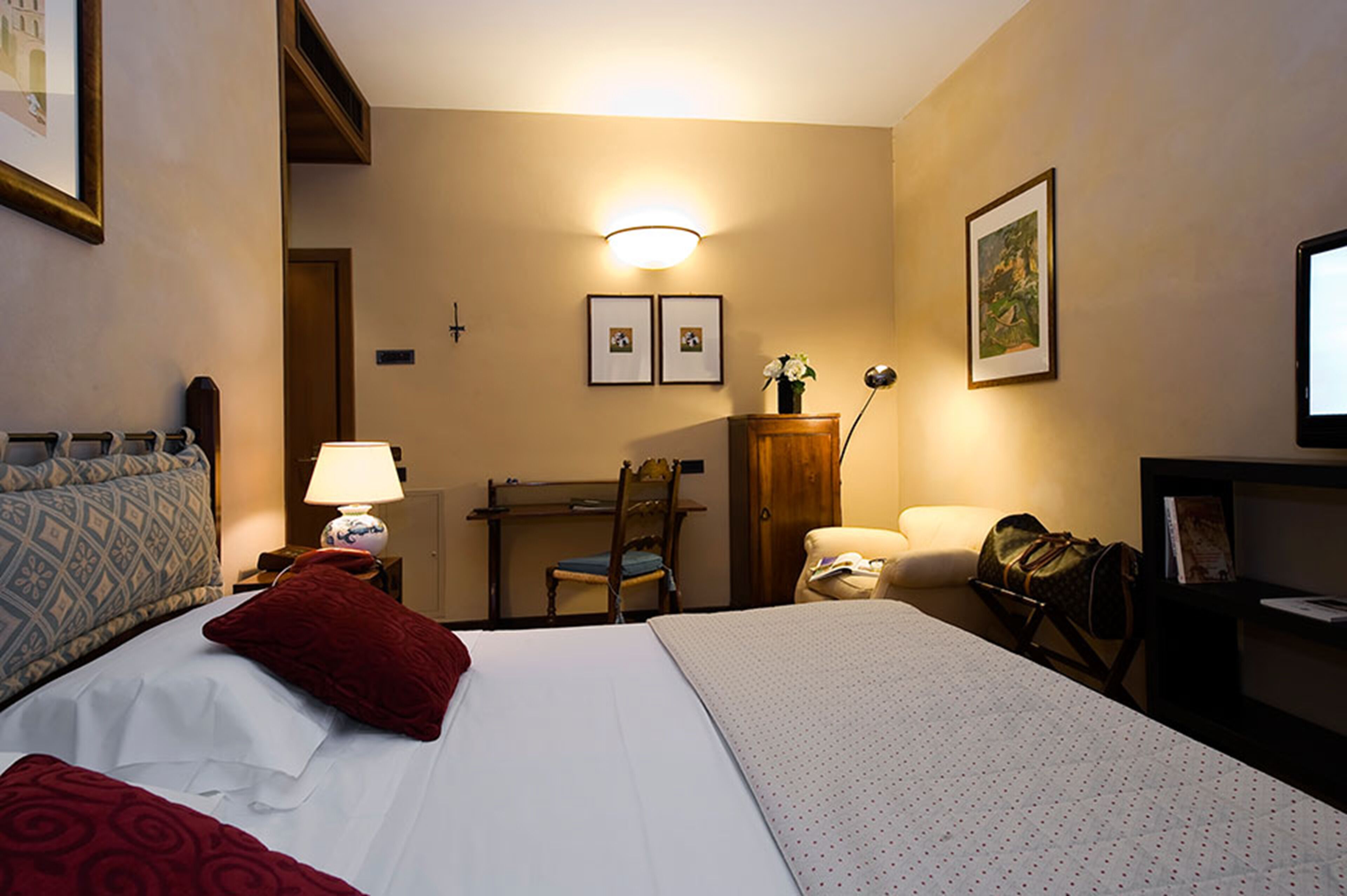 A hotel room with a bed, a desk, a chair, a lamp, a TV, and framed pictures on the wall.