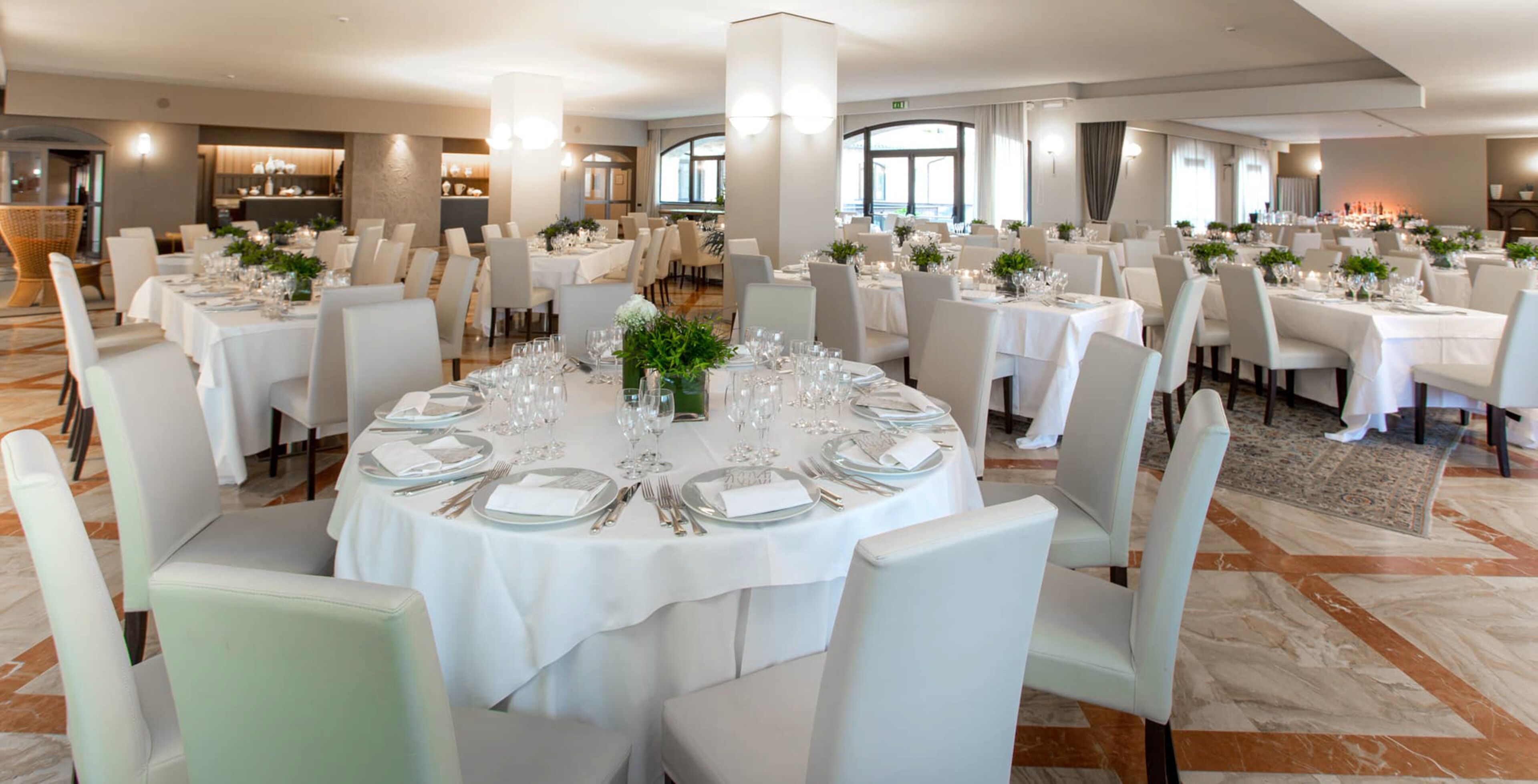 A spacious dining area with round tables set for multiple guests, featuring white tablecloths, neatly arranged cutlery, and small floral centerpieces.