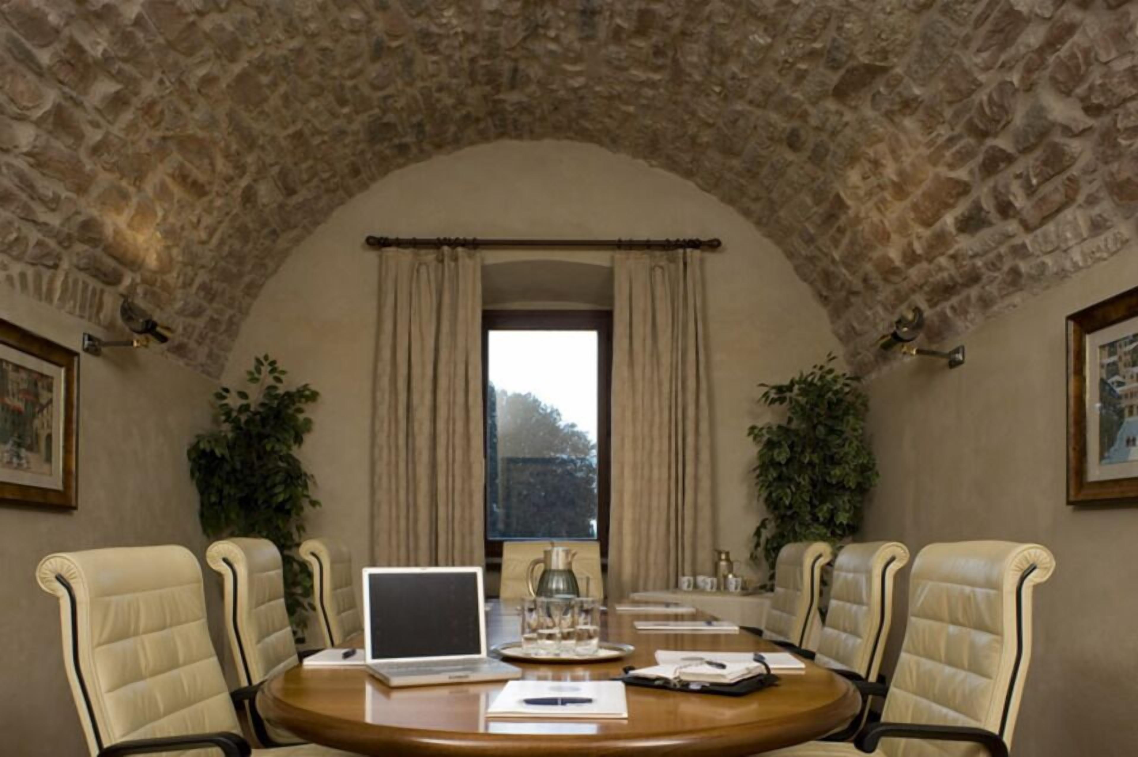 A conference room with a stone ceiling, a round table, beige chairs, a laptop, a pitcher, and a window with curtains.