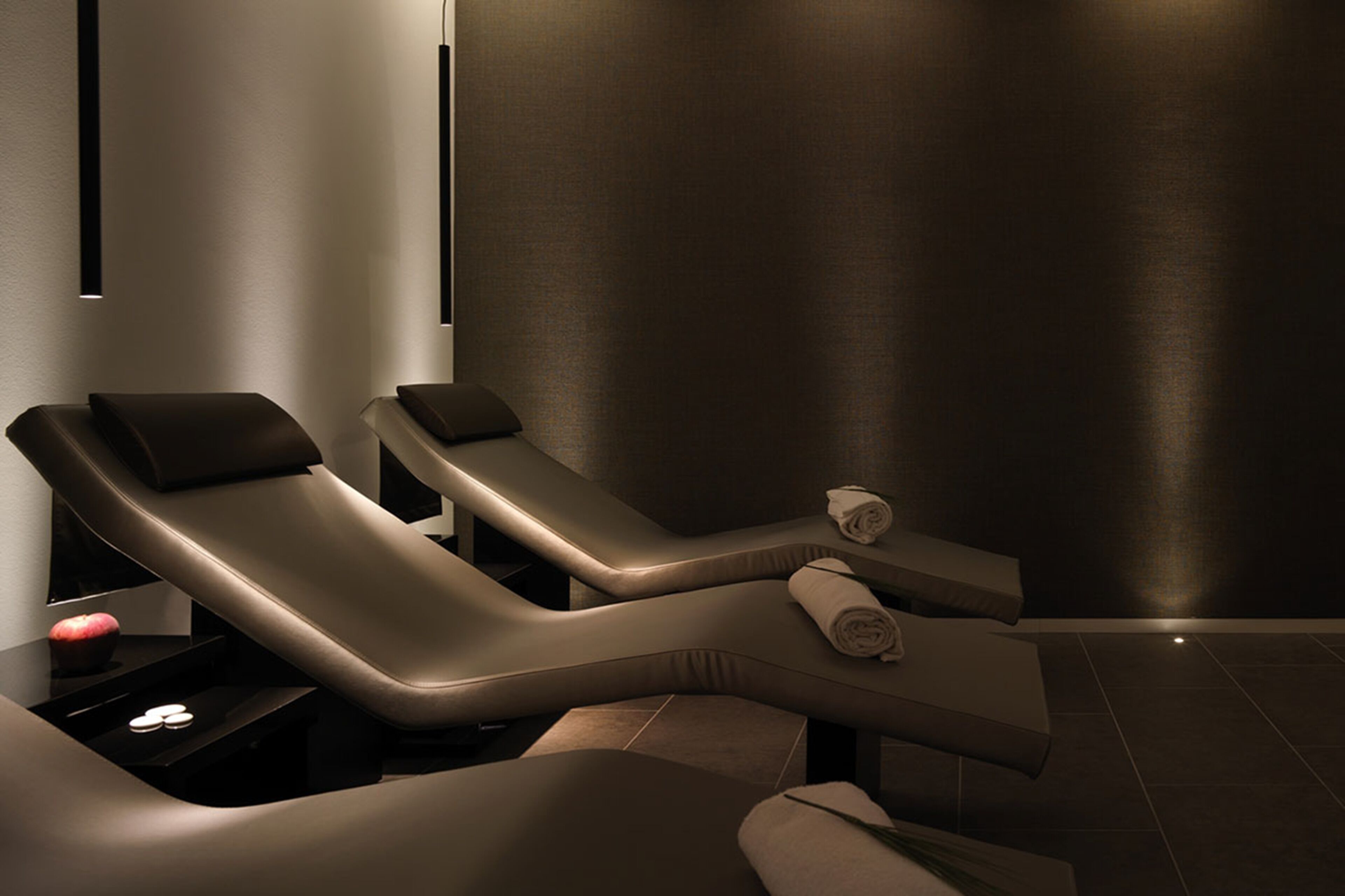A modern spa room with two reclining chairs, a small table with a towel and an apple, and a wall-mounted light fixture.