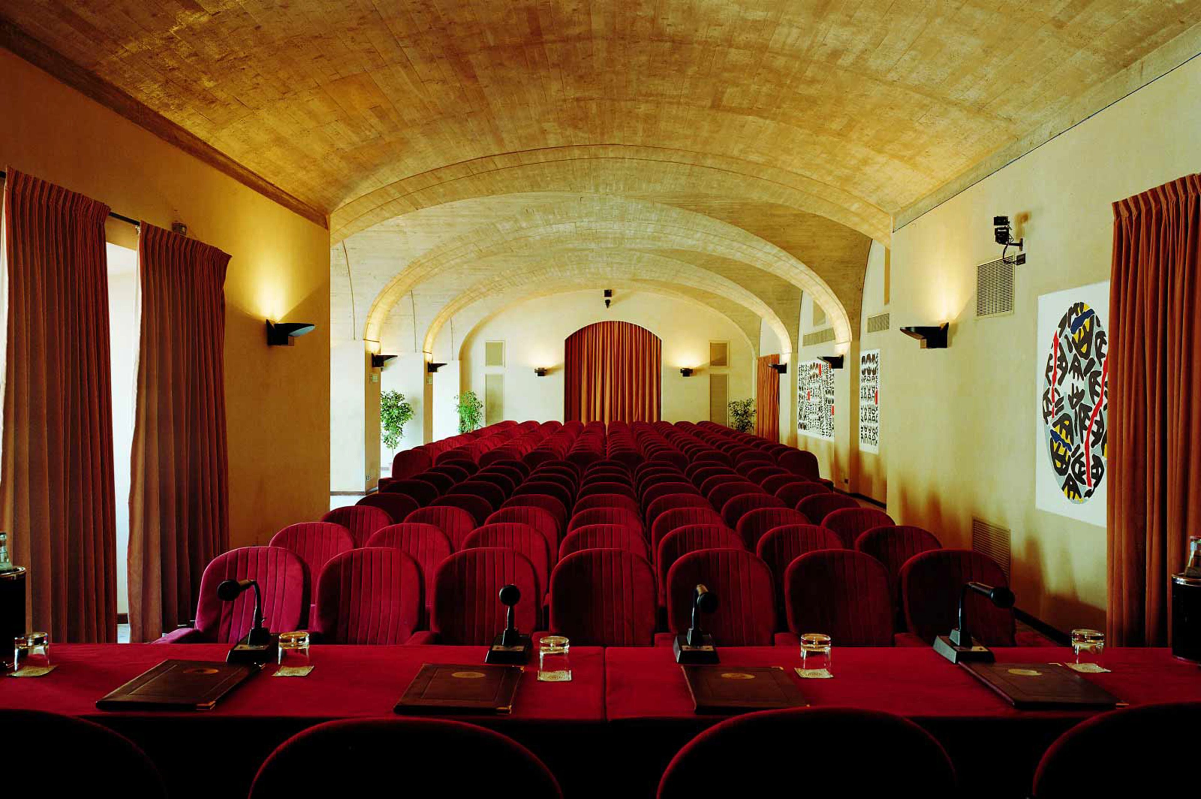 A conference room with rows of red chairs, a long table with microphones, and a high arched ceiling.