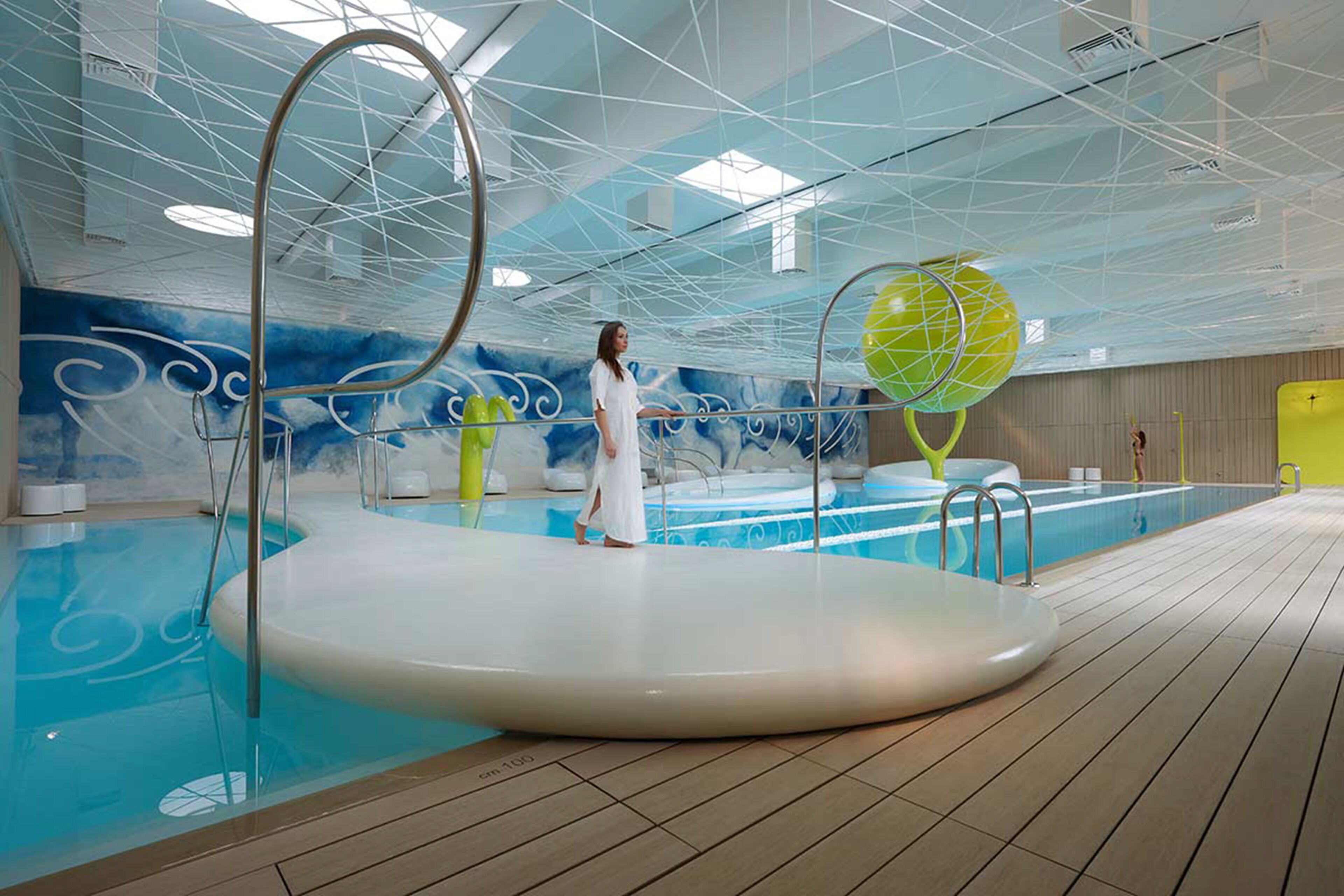 Une piscine intérieure moderne dotée d’un plafond au design unique, une personne se tenant près de la piscine et une terrasse en bois entourant l’espace.
