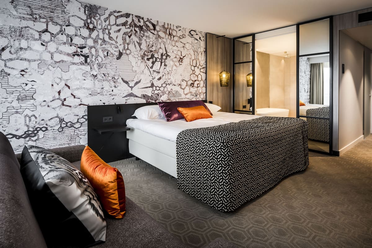 Een moderne hotelkamer met een groot bed, een bank en een gestreept voetenbankje. Er is een grote spiegel en een muur met een uniek abstract ontwerp.