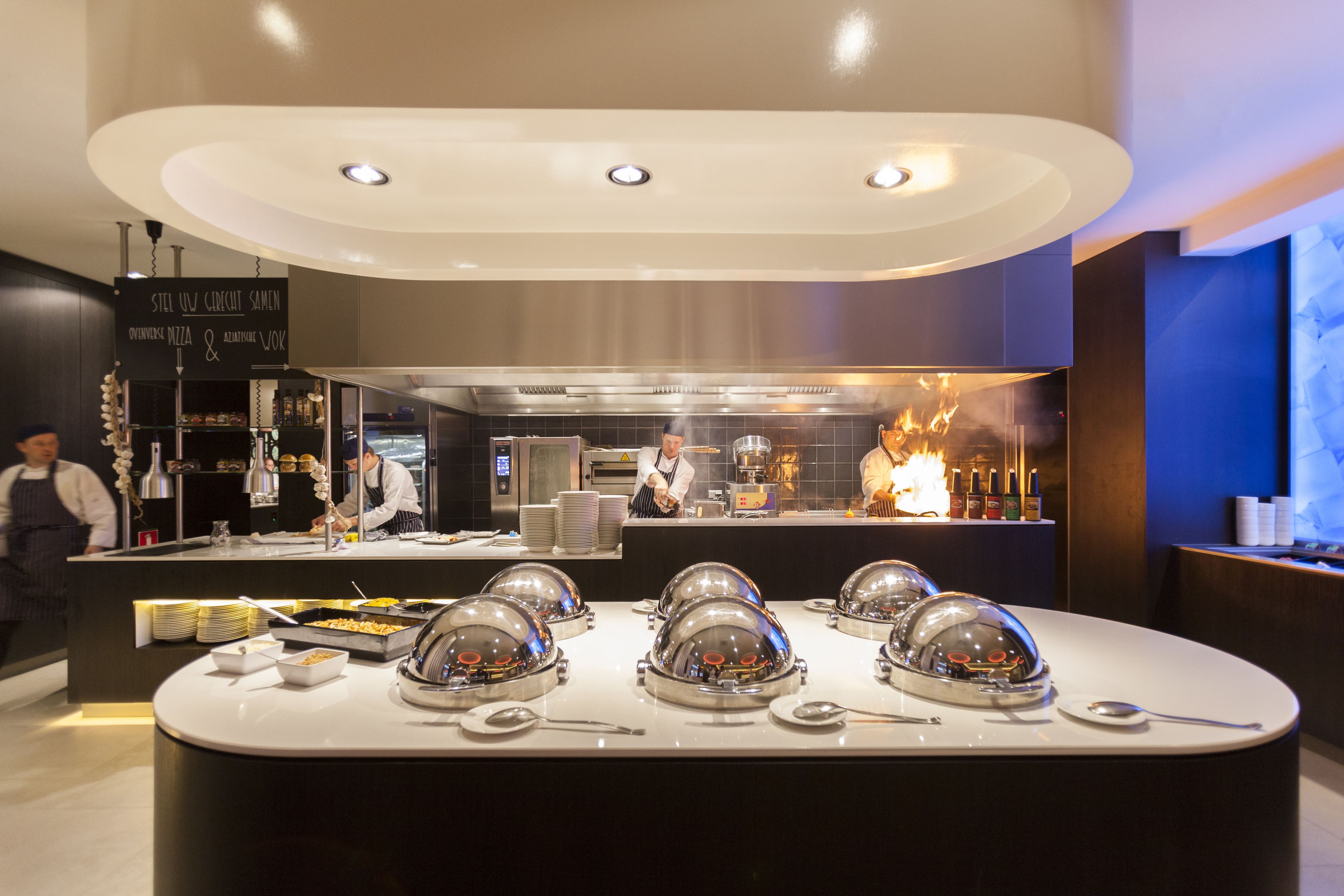 A modern restaurant with a central counter, buffet area, and a visible kitchen with chefs.