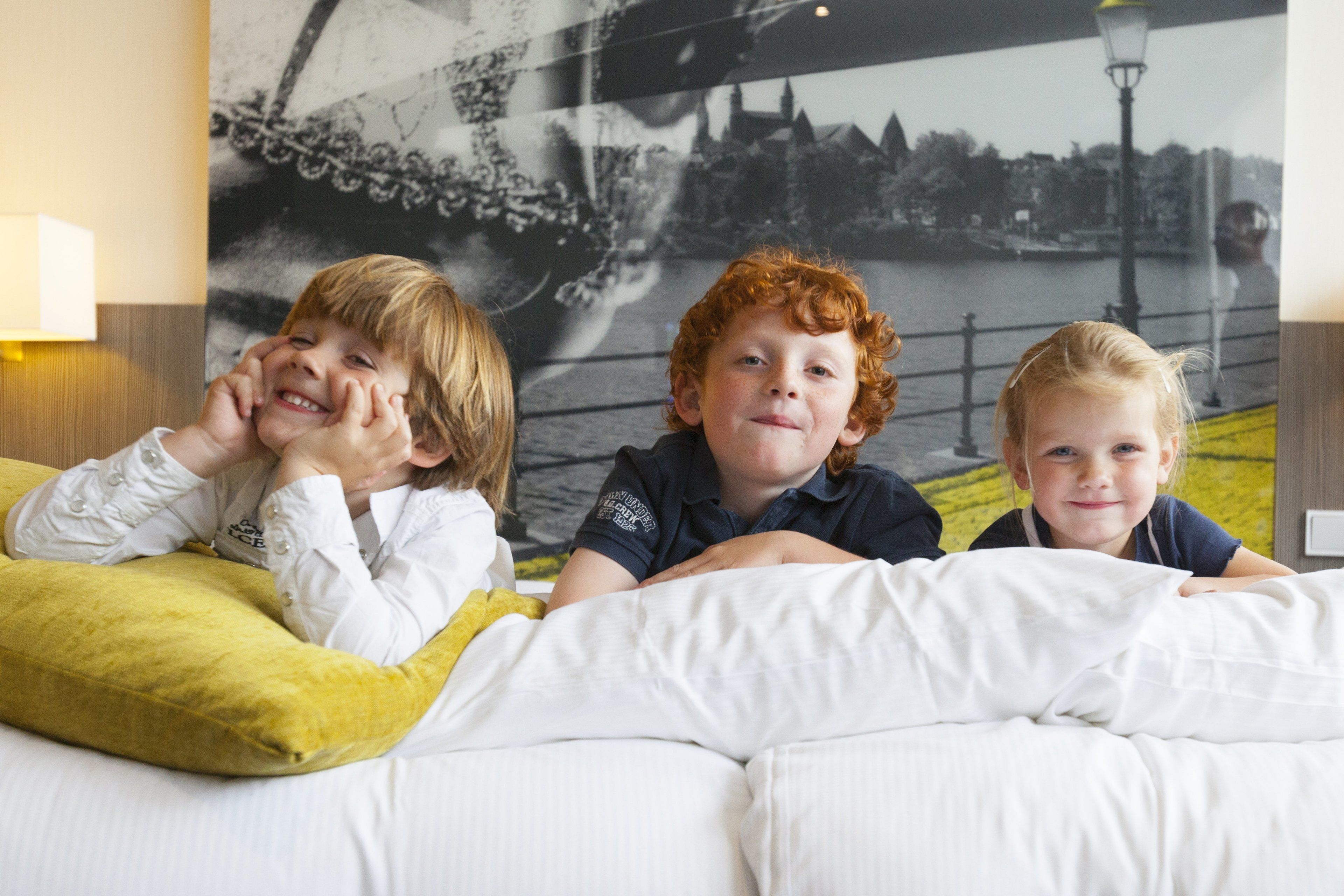 Three children lying on a bed with a cityscape mural in the background.