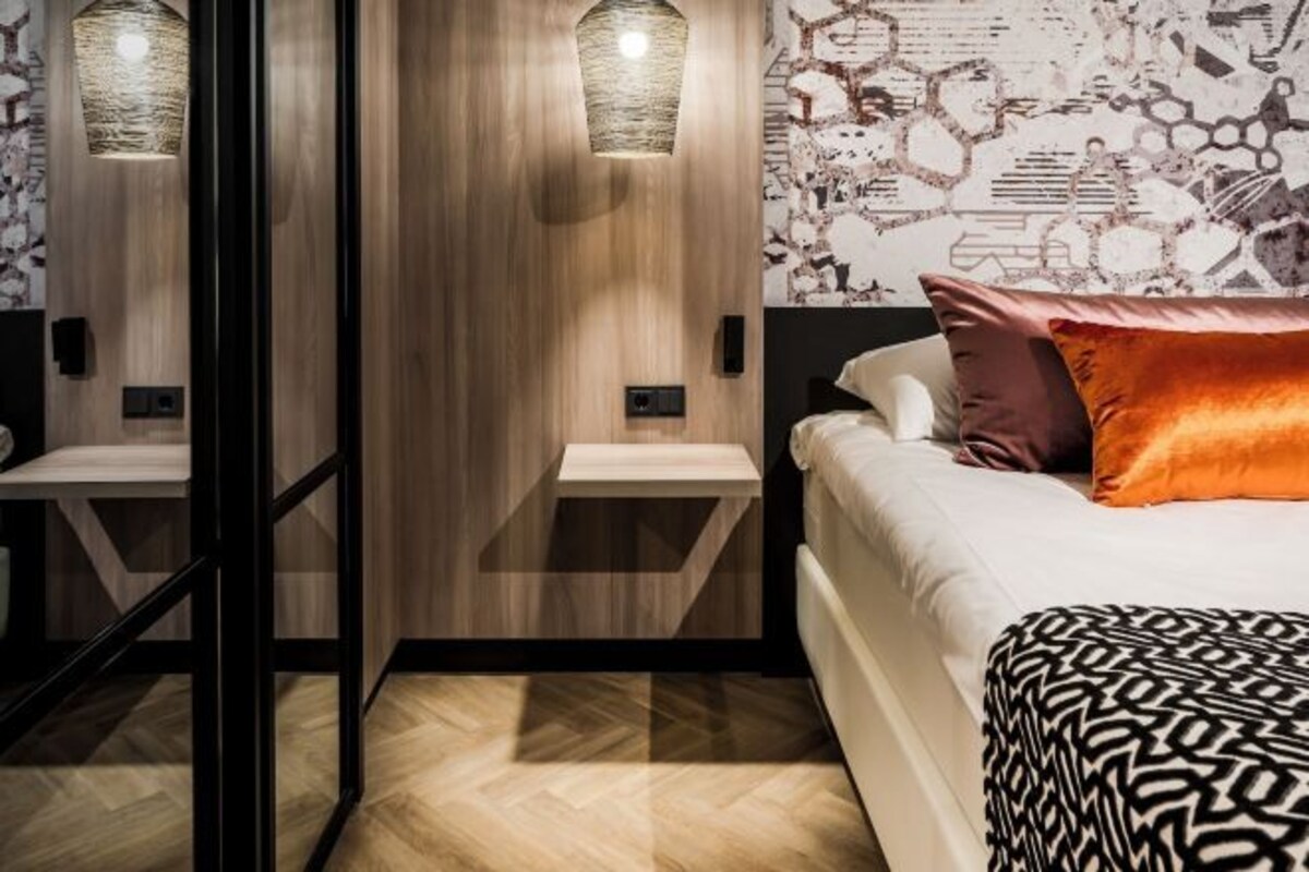 A modern hotel room with a bed, a nightstand, a wall-mounted lamp, and a patterned wall.