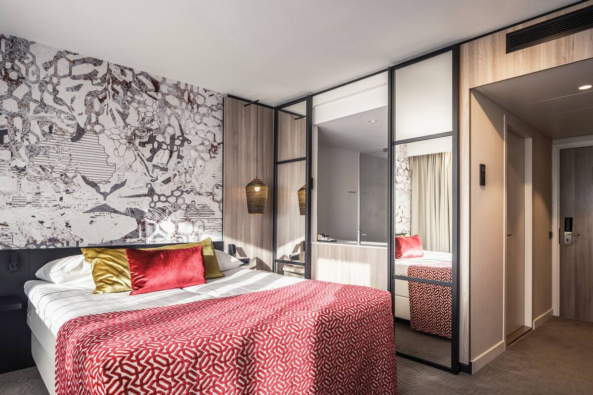 Een moderne hotelkamer met een groot bed, een spiegel en uitzicht op de stad door grote glazen deuren.