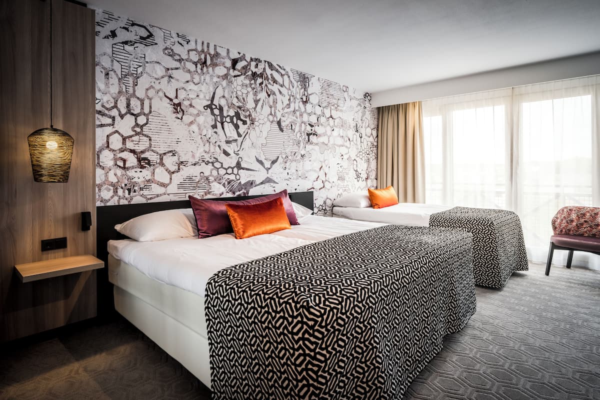 Een hotelkamer met twee bedden, een behang met patroon, een houten kastje, een nachtkastje en een raam met gordijnen.