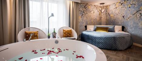 A bedroom with a large bed, a sitting area with chairs, a bathtub with rose petals, and a wall with a floral mural.