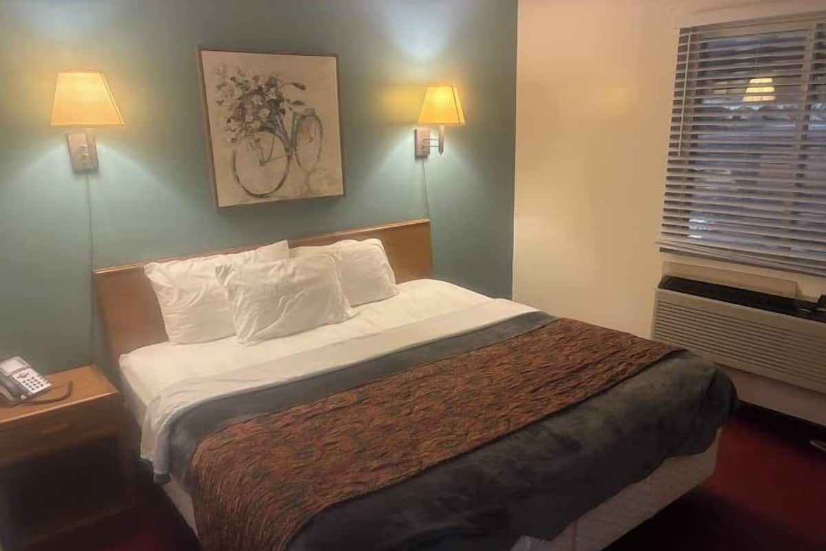 A hotel room with a bed, two bedside lamps, a telephone, a painting, and a window with blinds.