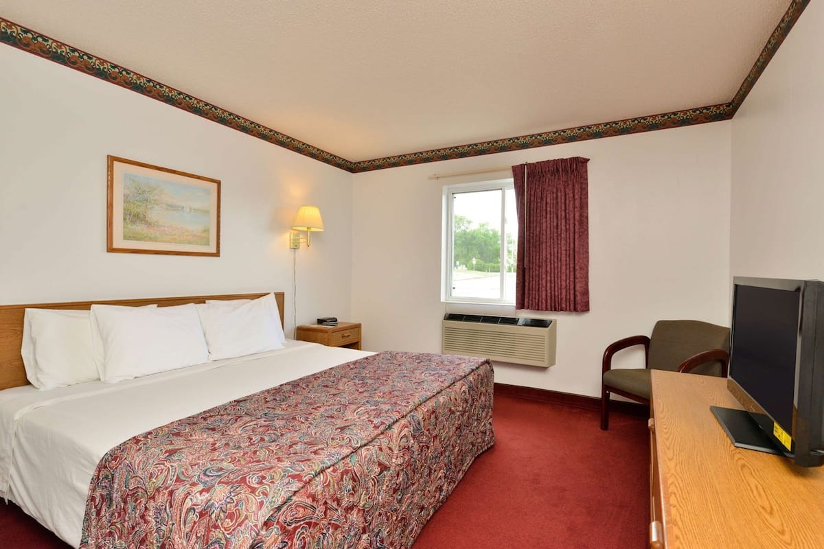A hotel room with a bed, a desk with a television, a window with curtains, a wall-mounted air conditioner, and a painting on the wall.