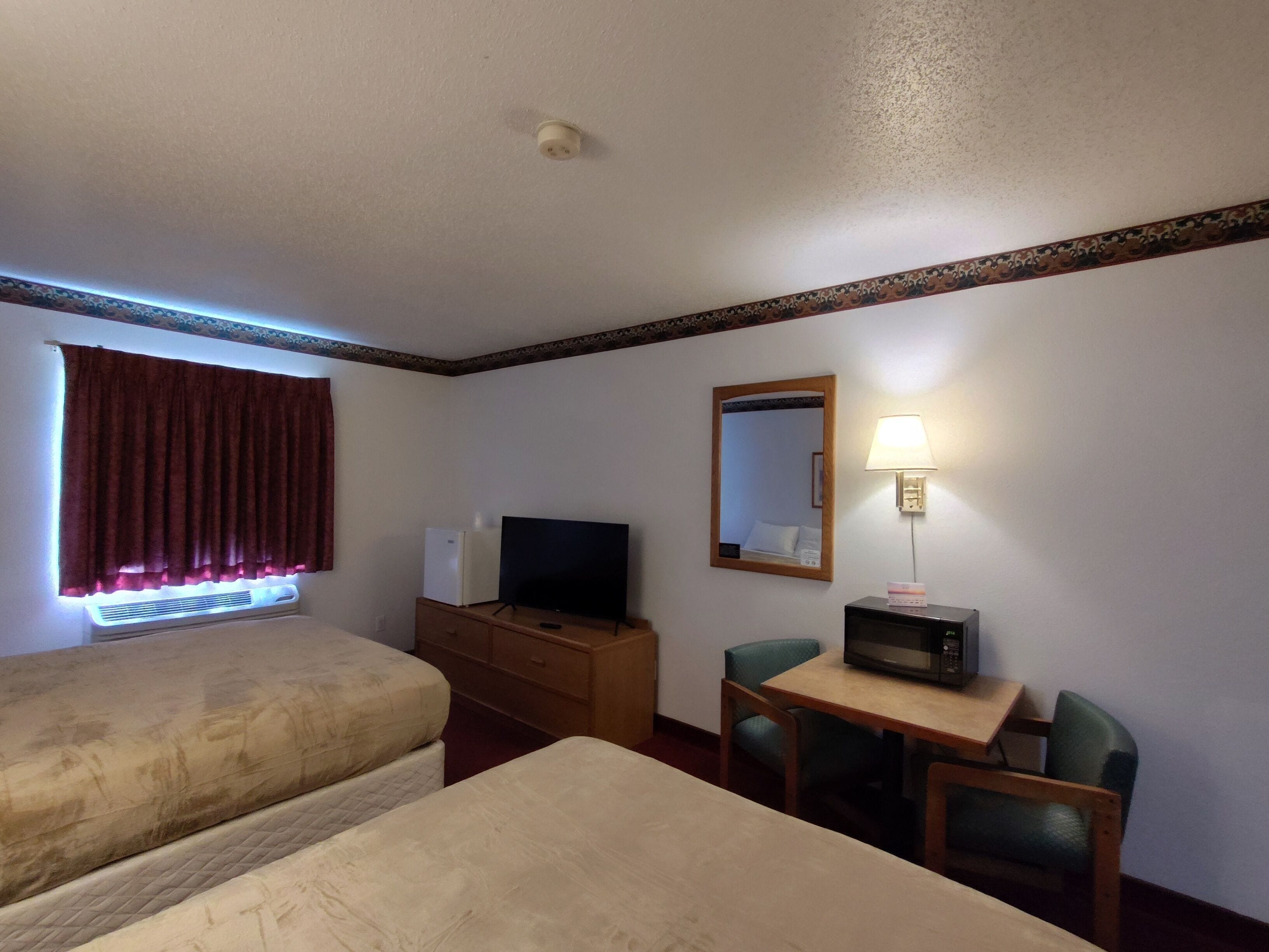 A hotel room with two beds, a desk with a television, a microwave, a lamp, and a window with curtains.