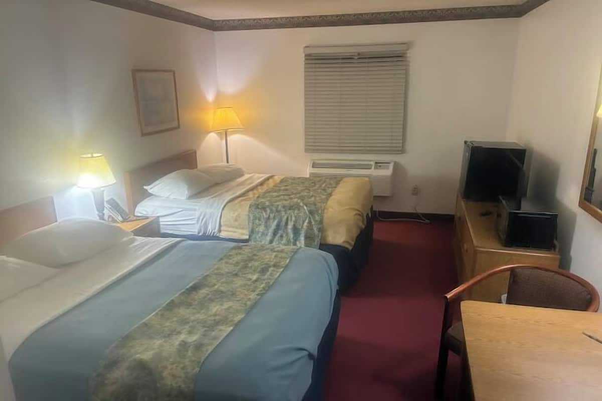 A hotel room with two beds, a desk, a chair, a lamp, an air conditioner, and a television.