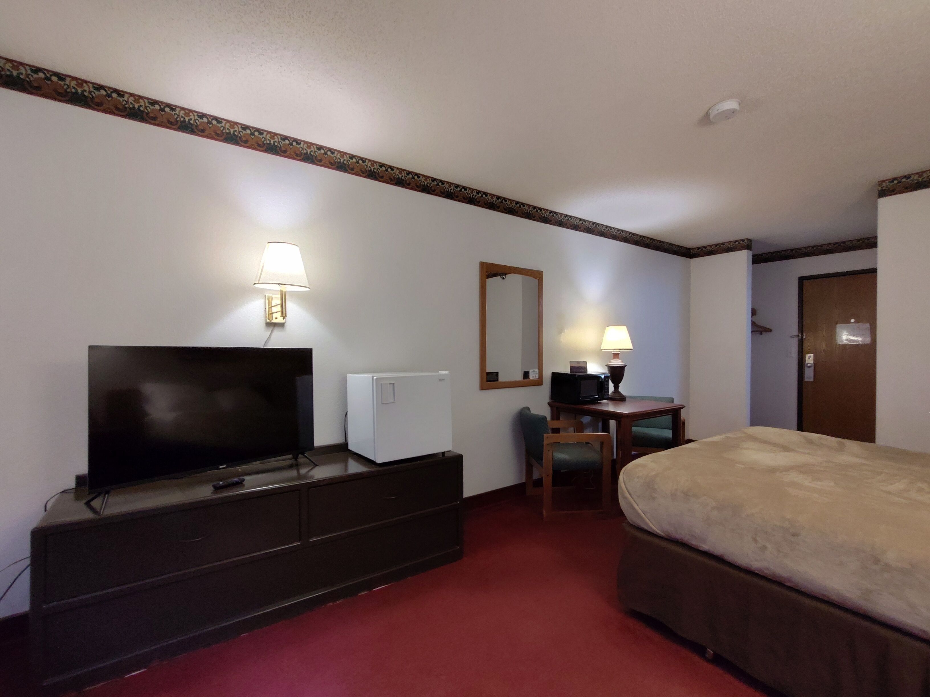 A hotel room with a bed, a TV, a desk with a lamp, a mirror, and a refrigerator.