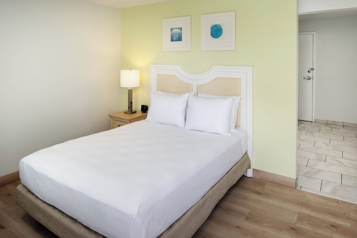Una cama bien hecha con un cabecero blanco, una lámpara de noche y dos cuadros en la pared.