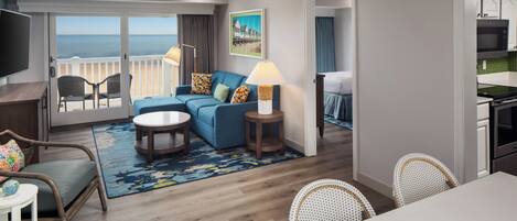 A modern hotel room with a balcony, a sofa, a dining table, and a kitchen area.