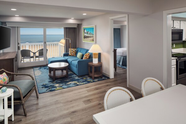 A modern hotel room with a balcony, a sofa, a dining table, and a kitchen area.