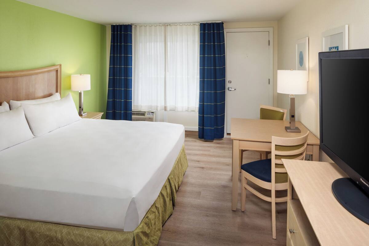 Habitación de hotel con cama, escritorio, silla, televisor y ventana con cortinas.