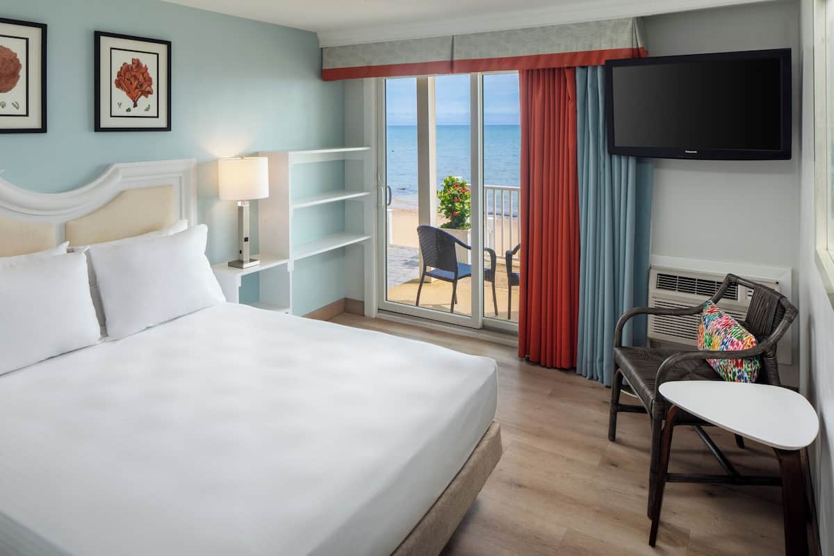 Habitación de hotel con cama, televisión, una silla, una mesita y vista al océano a través de una puerta corrediza de vidrio.