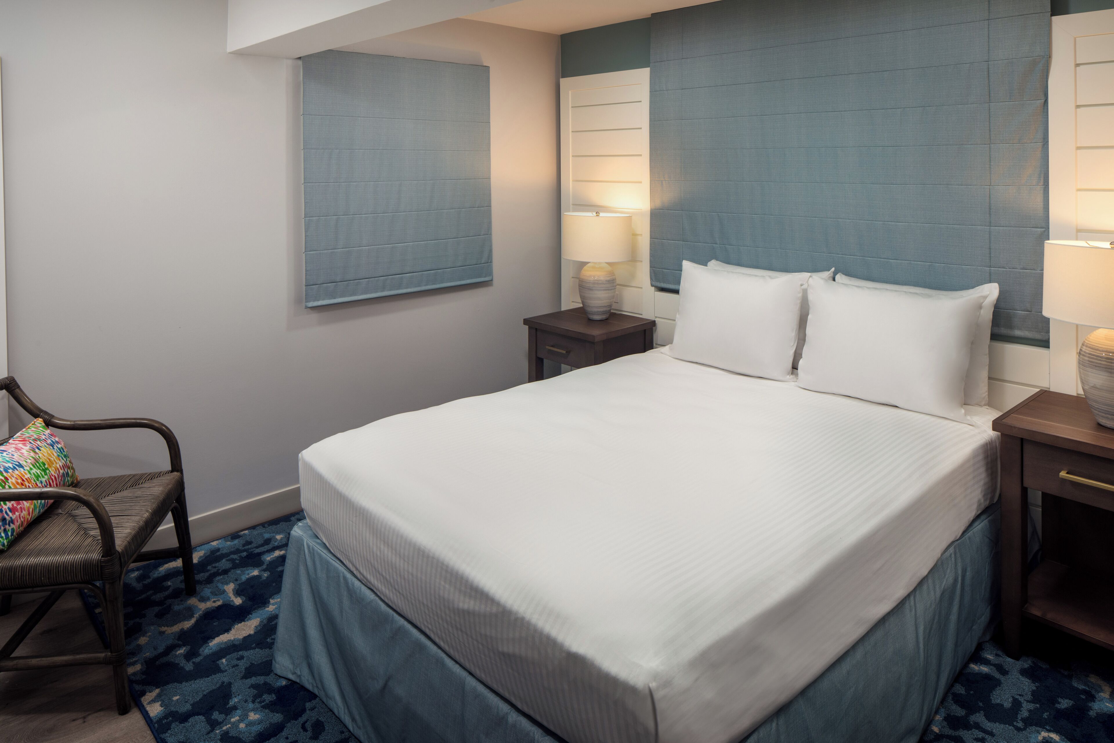 A neatly made bed with white linens, two bedside lamps, and a chair with a colorful cushion.