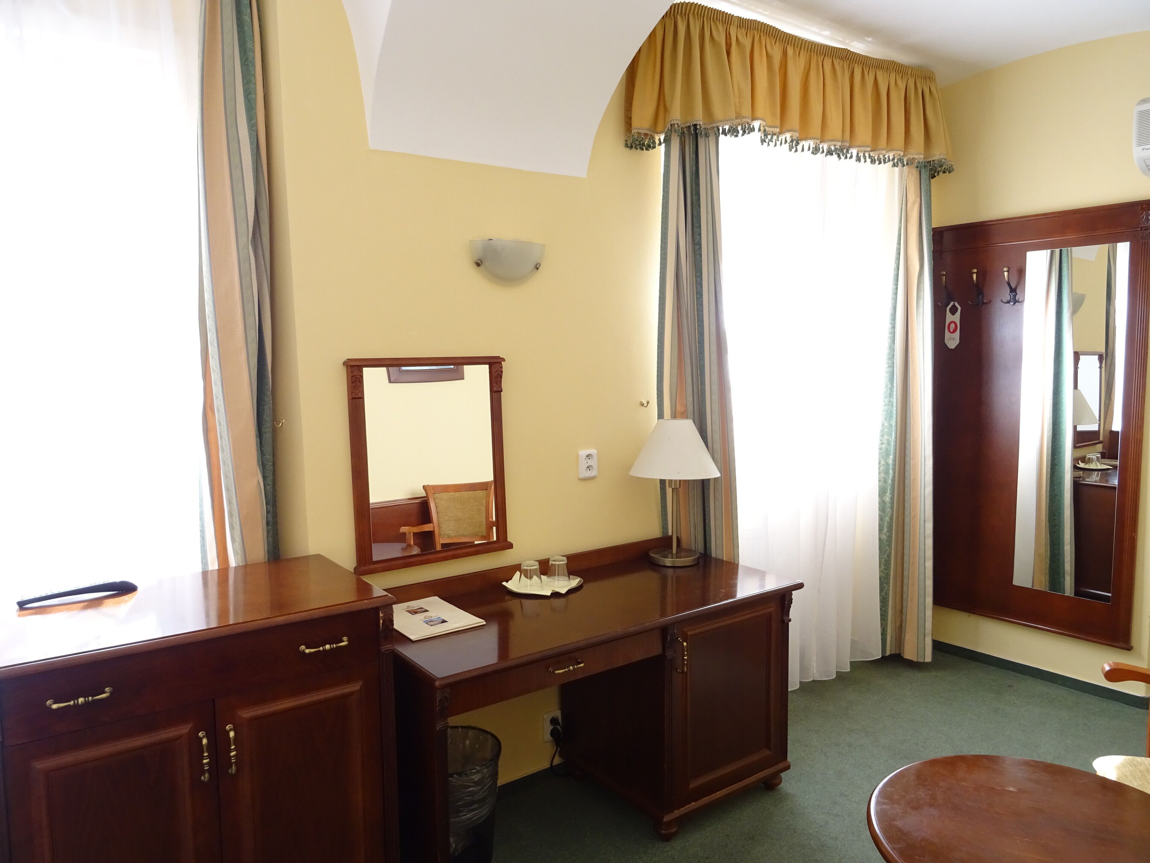 Quarto de hotel com escrivaninha de madeira, espelho, luminária, cadeira e porta com brasão.