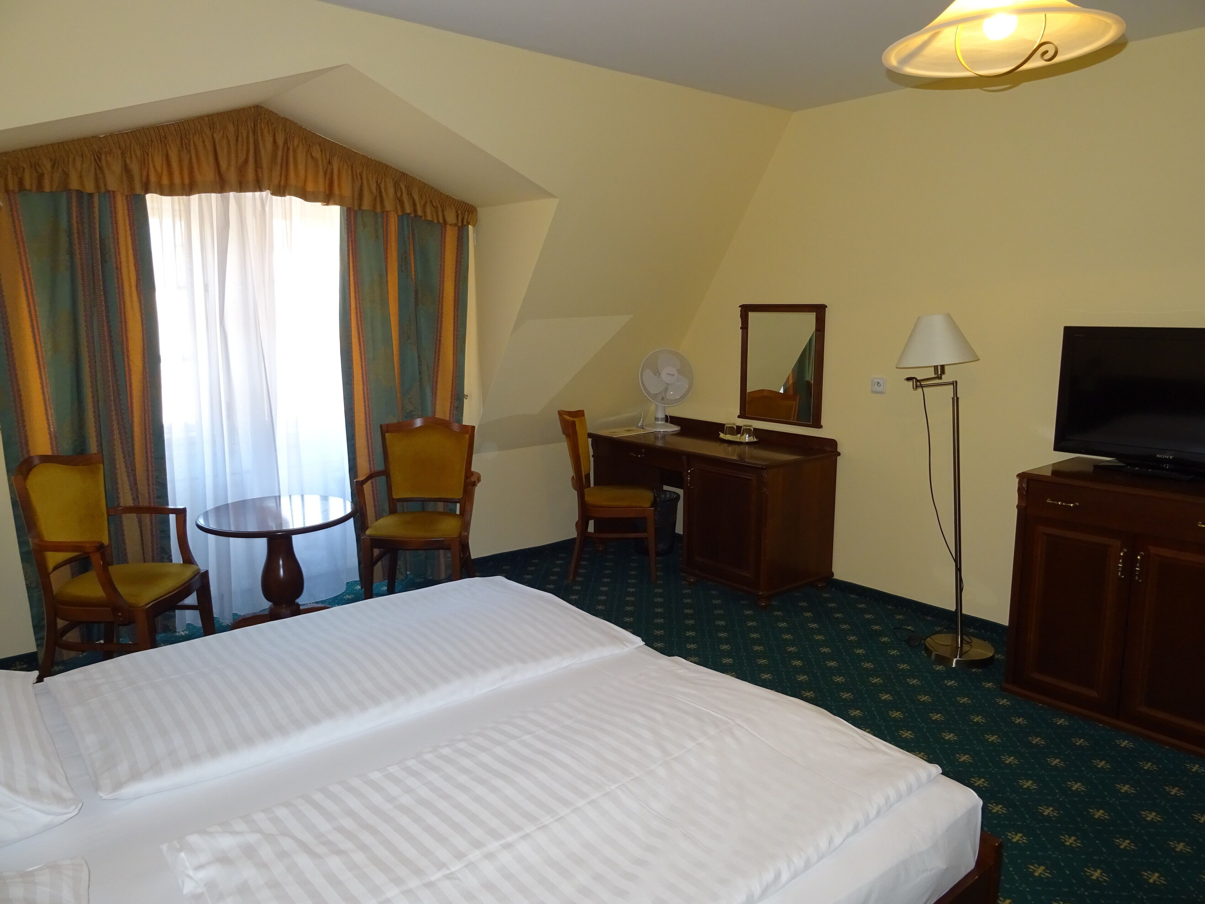 Quarto de hotel com uma cama grande, uma escrivaninha com cadeira, televisão e uma luminária.
