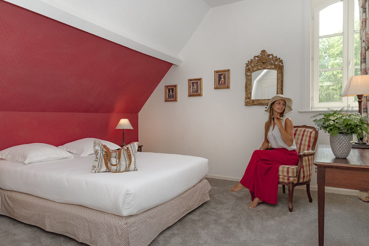 A hotel room with a large bed, a red accent wall, a mirror, and a window with a view of greenery.
