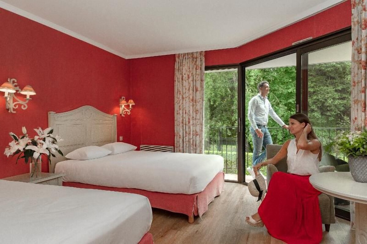 A hotel room with two beds, a balcony, and a view of greenery.