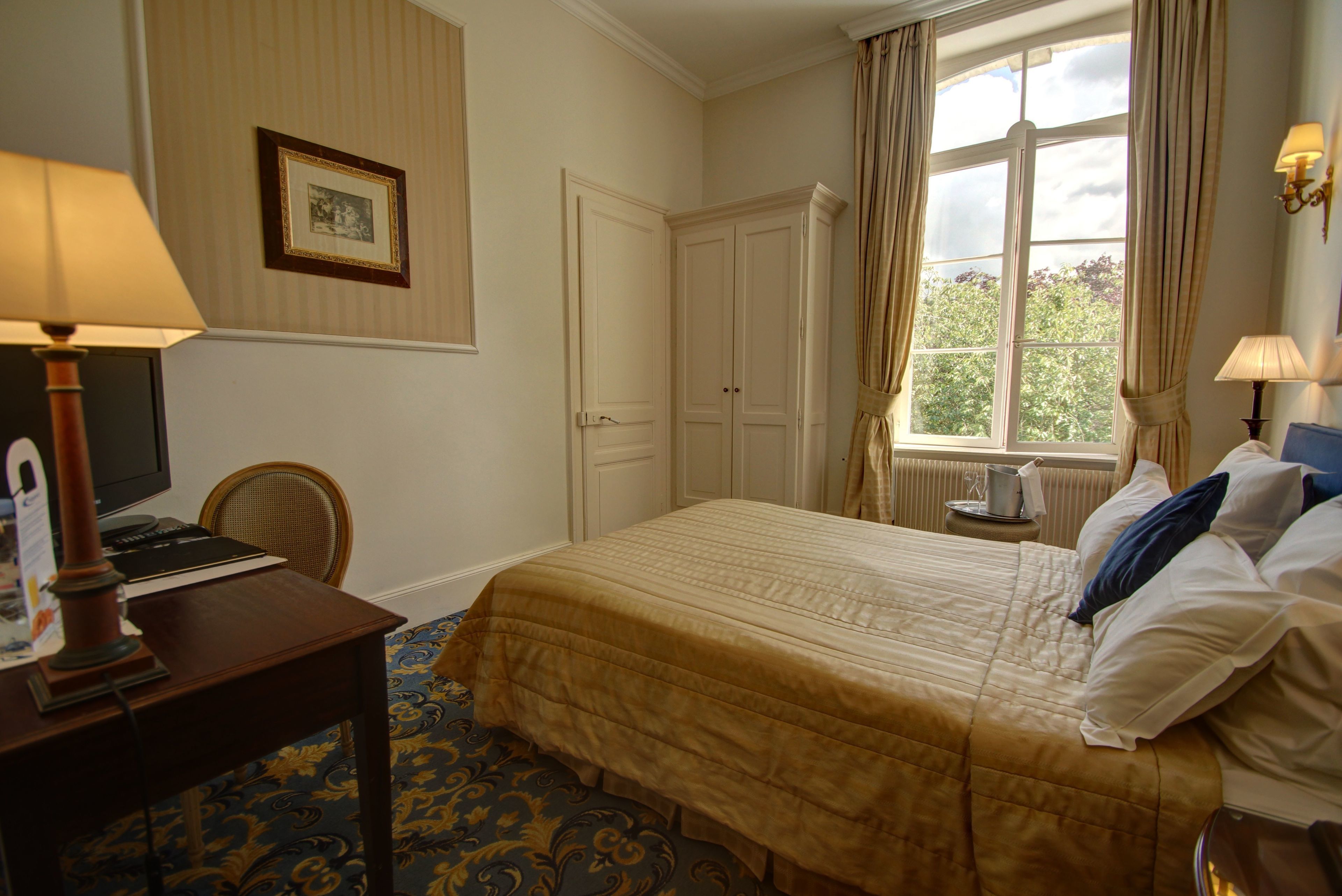 A hotel room with a bed, a desk with a lamp, a chair, a framed picture on the wall, and a window with curtains.