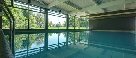 An indoor swimming pool with clear glass walls, a metal roof, and a view of trees outside.