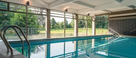Una piscina interior con paredes de vidrio transparente, barandillas de metal y vistas a un paisaje verde.