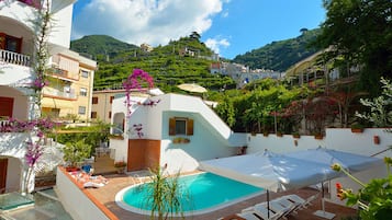 Area piscina con vasca azzurra e cristallina, circondata da pareti bianche e un solarium in legno. Sono presenti lettini e ombrelloni per l'ombra.