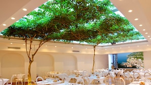 Um restaurante com mesas redondas, cadeiras brancas e uma grande árvore ao centro.