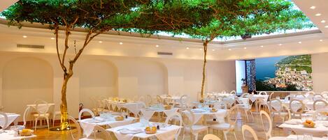 A restaurant with round tables, white chairs, and a large tree in the center.