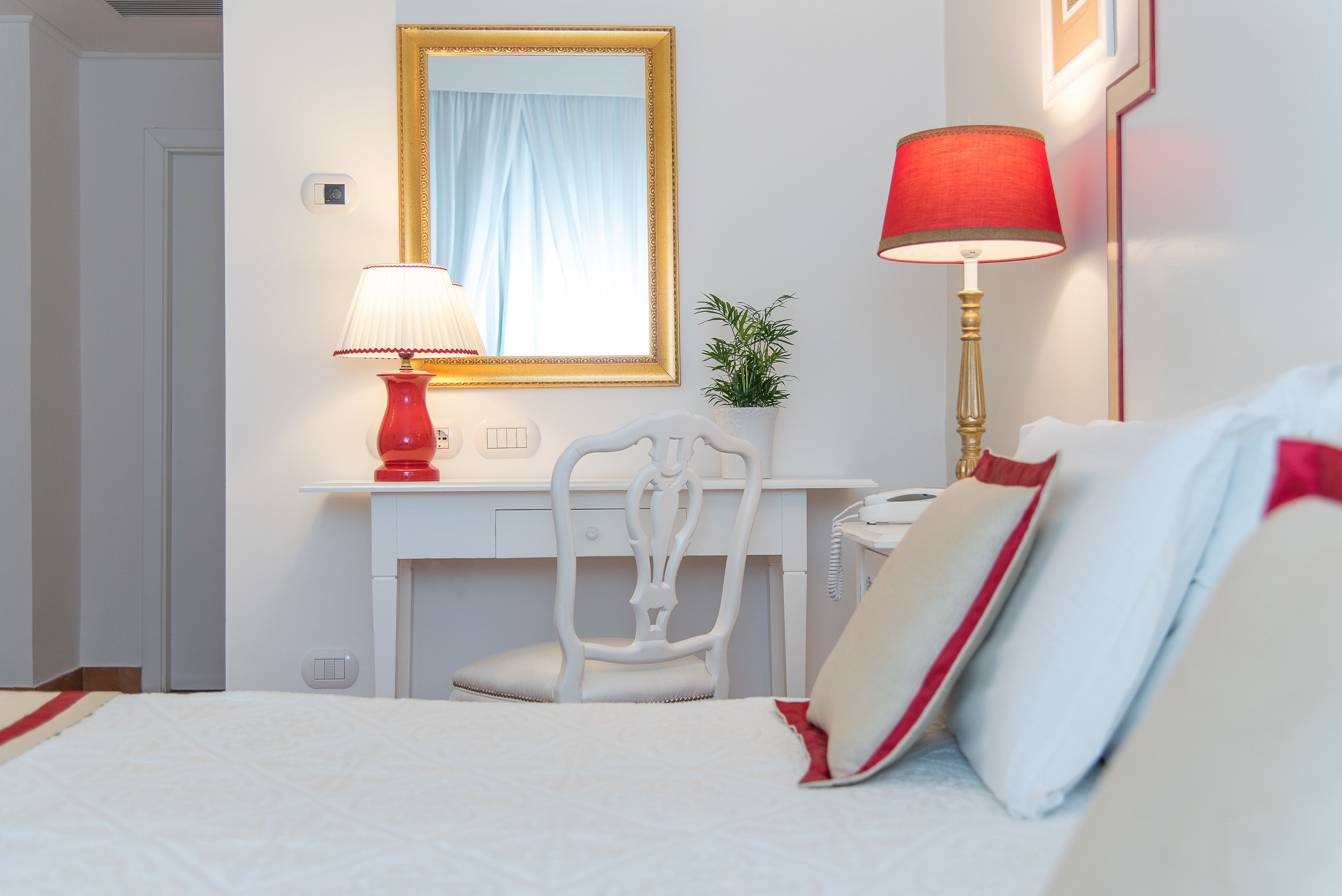 A hotel room with a bed, a desk with a chair, a telephone, a lamp, a mirror, and a small plant.