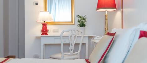 A hotel room with a bed, a desk with a chair, a telephone, a lamp, a mirror, and a small plant.