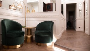 Um corredor com duas poltronas de veludo verde e uma mesa dourada, paredes brancas e chão de madeira.