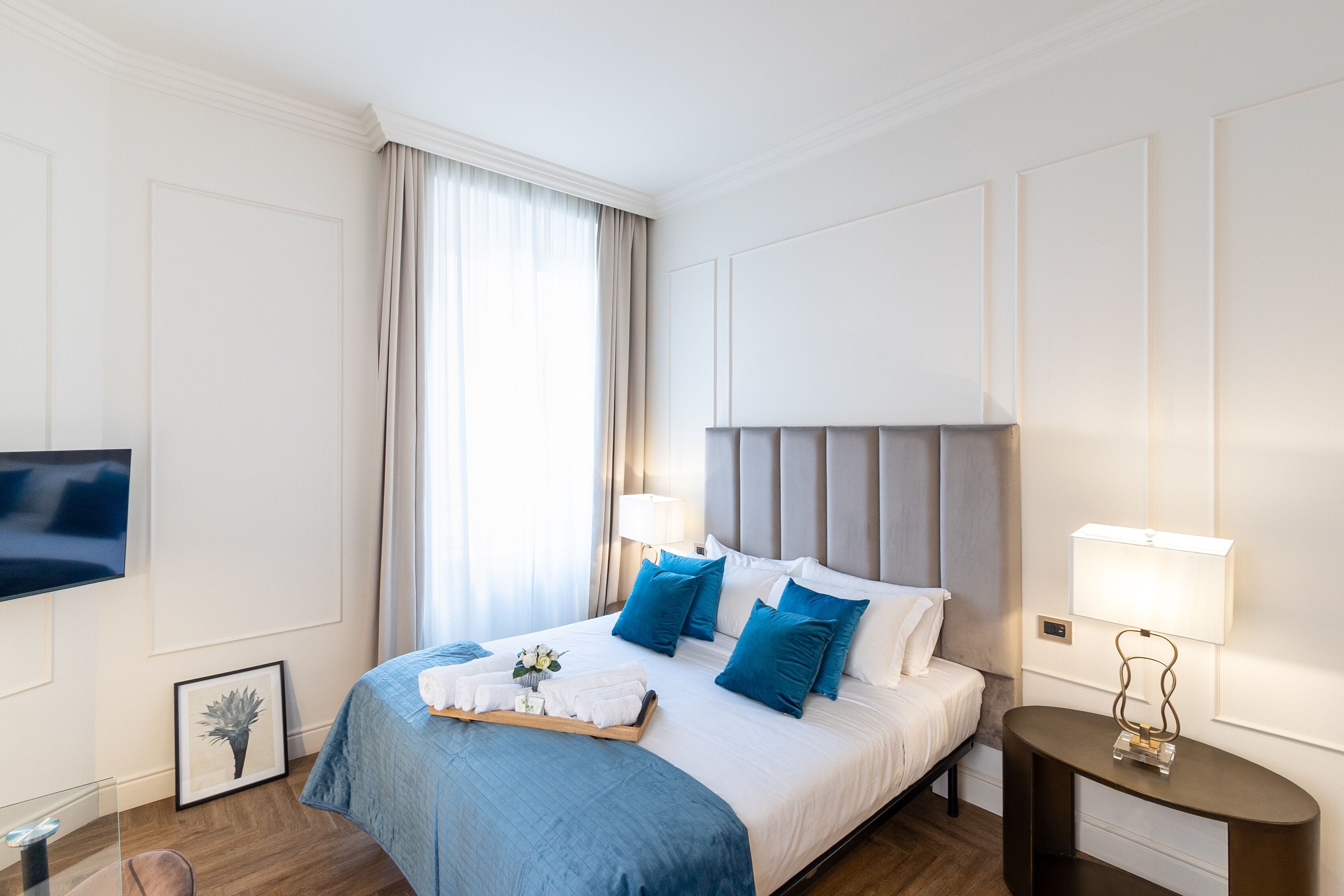 A hotel room with a large bed, a bedside table with a lamp, a flat-screen TV mounted on the wall, and a framed picture on the floor.