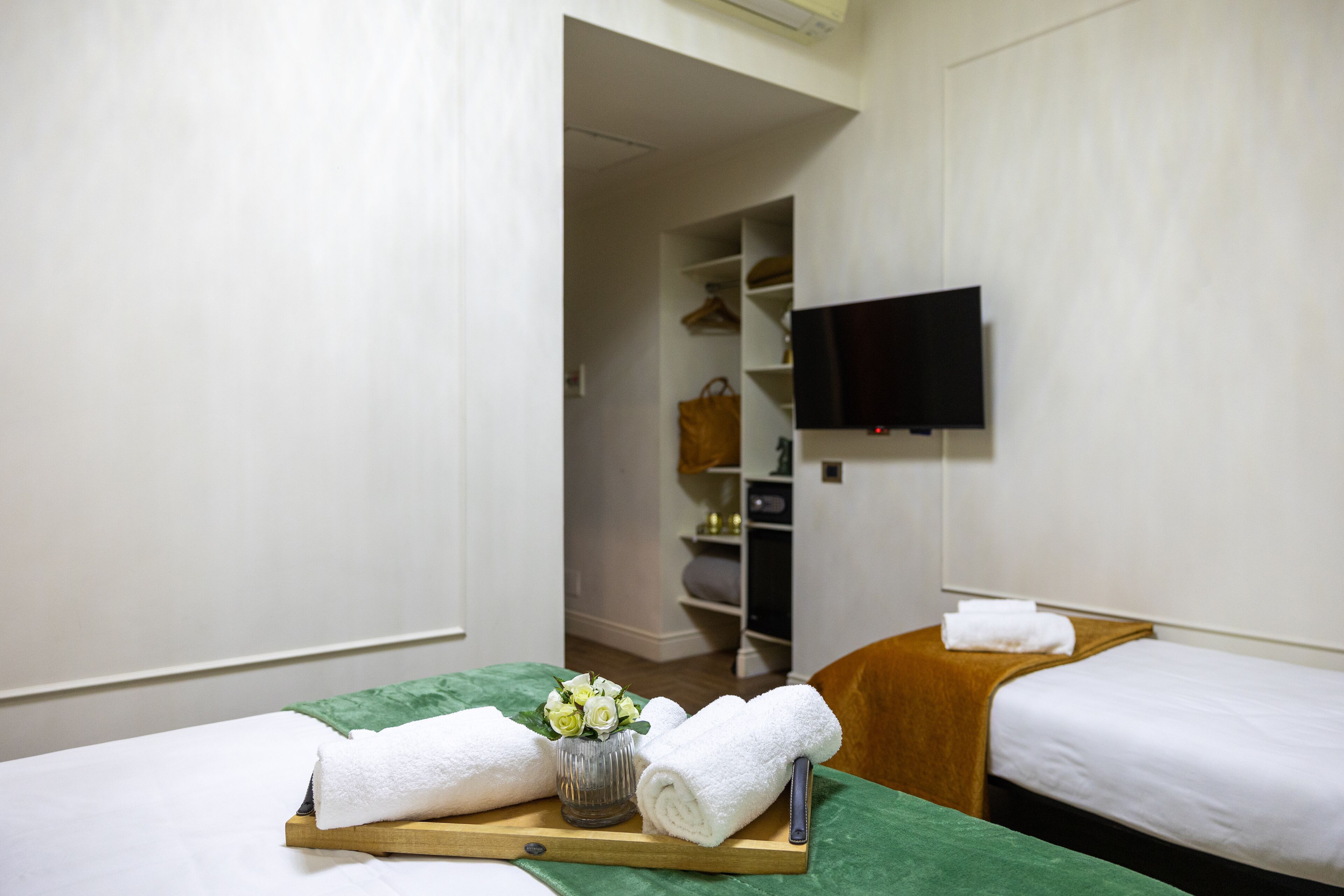 Habitación de hotel con dos camas, un televisor montado en la pared, una mesita de madera con toallas y un jarrón de flores, y un armario con ropa colgada.