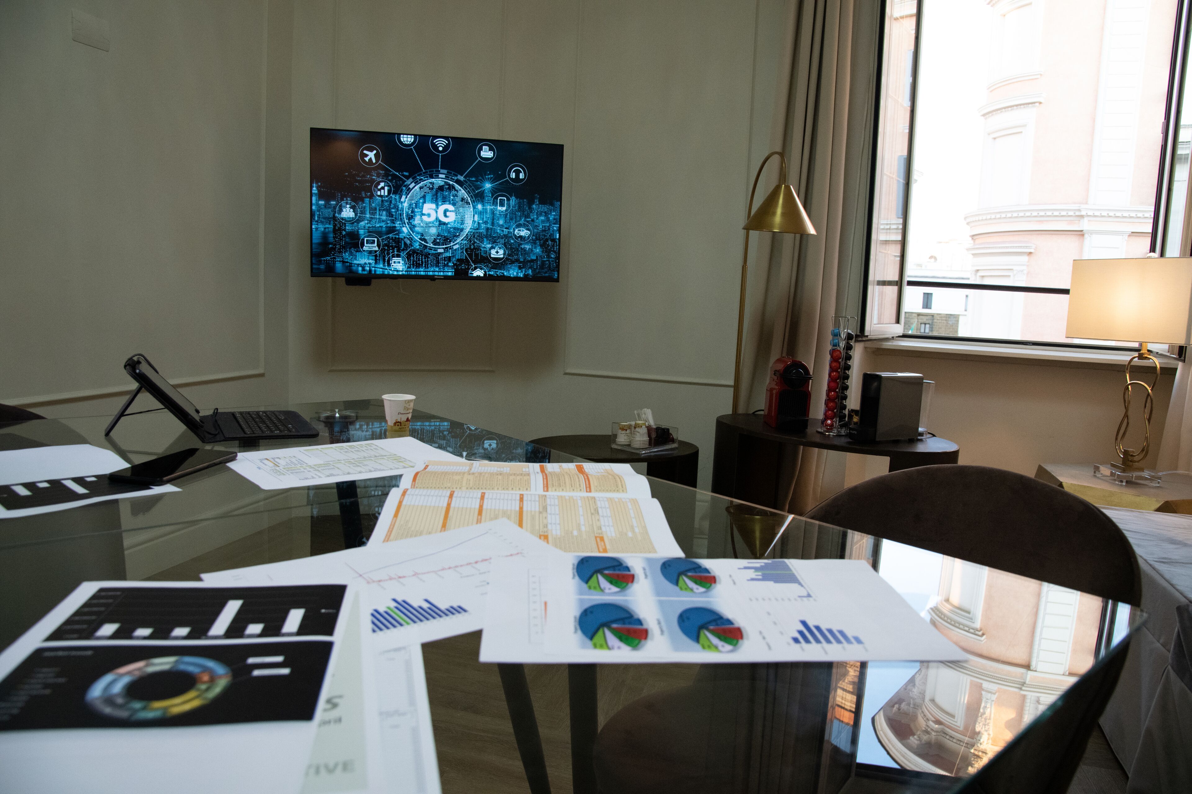Une chambre d’hôtel équipée d’un bureau, de documents, d’un ordinateur portable, d’un téléviseur affichant un réseau 5G et offrant une vue sur les bâtiments par la fenêtre.