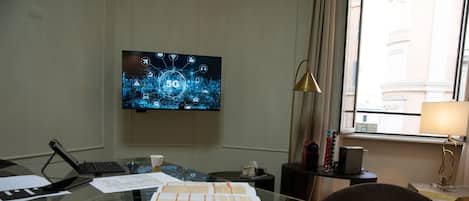 A hotel room with a desk, papers, a laptop, a TV displaying a 5G network, and a view of buildings through the window.