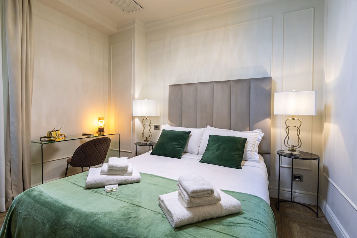 Ein Hotelzimmer mit einem ordentlich bezogenen Bett, grüner Bettwäsche, zwei Nachttischlampen, einem kleinen Tisch mit Lampe und einem Stuhl.